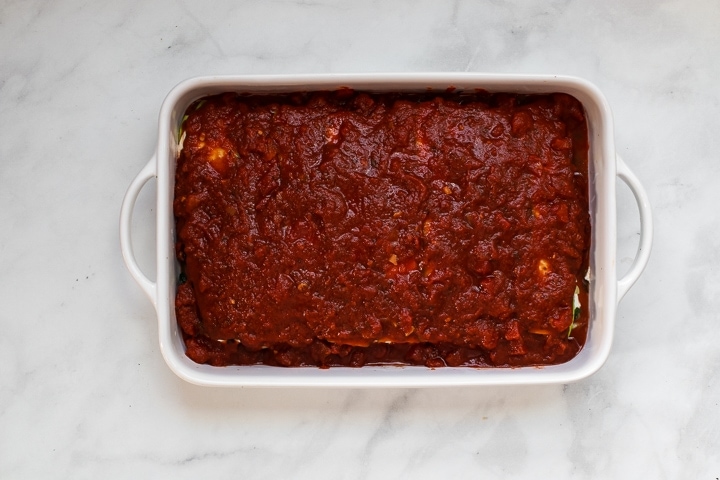 White baking dish with top layer of zucchini slices and tomato sauce spread over the lasagna on gray marble. 