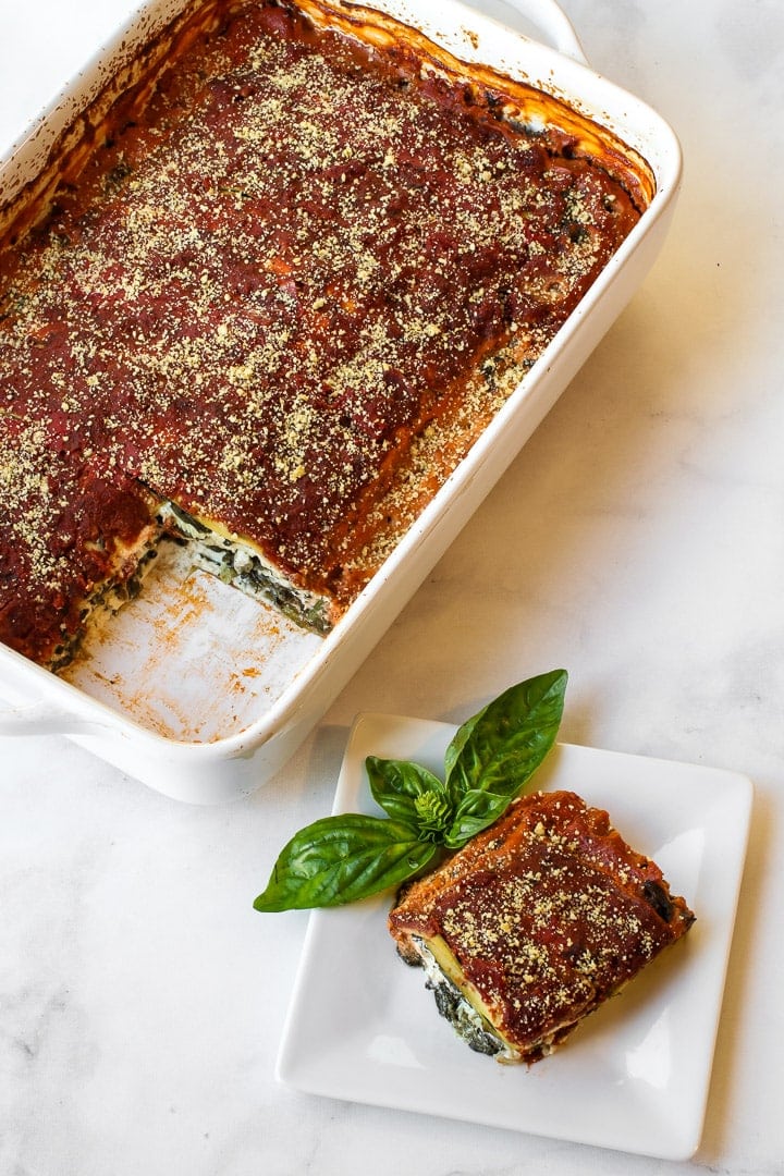 White baking dish with vegan Parmesan-topped baked lasagna, one piece cut and on a white plate with basil sprig, on gray marble.