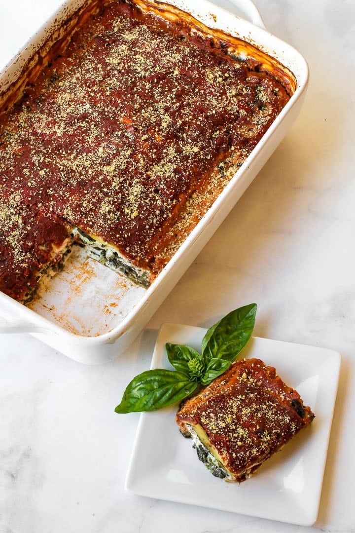 Overhead shot of white baking dish with vegan zucchini lasagna, one piece on white plate with basil sprig, on white marble.