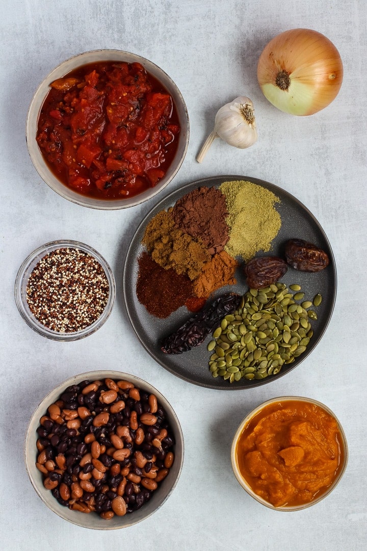 Pumpkin chile mole ingredients on a gray background: mole spices, pepita seeds, onion, garlic, black & pinto beans, tricolored quinoa, diced tomatoes, pumpkin puree.