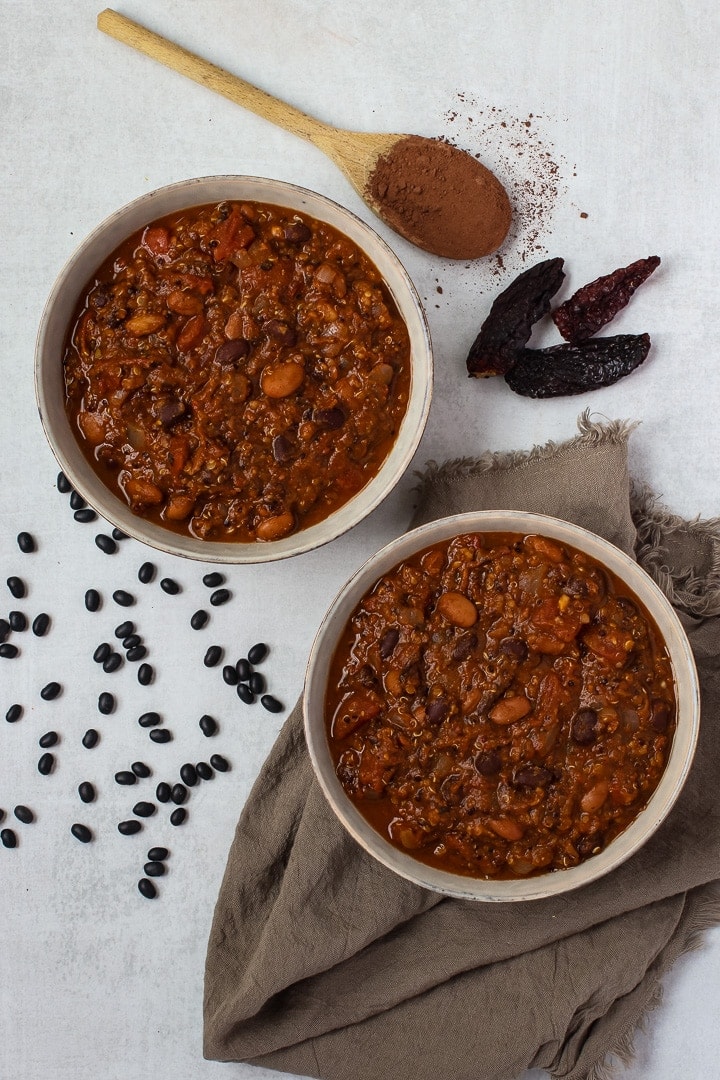 2 bowls of vegan pumpkin chili mole with on a brown towel, with dried black beans, a wooden spoon with cocoa powder and dried chilis.l