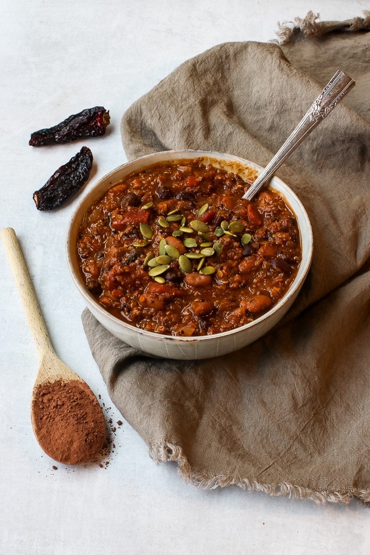 Bowl of pumpkin chili mole with pepita seeds and a spoon on a brown towel, with wooden spoon with cocoa powder and dried chilis.l