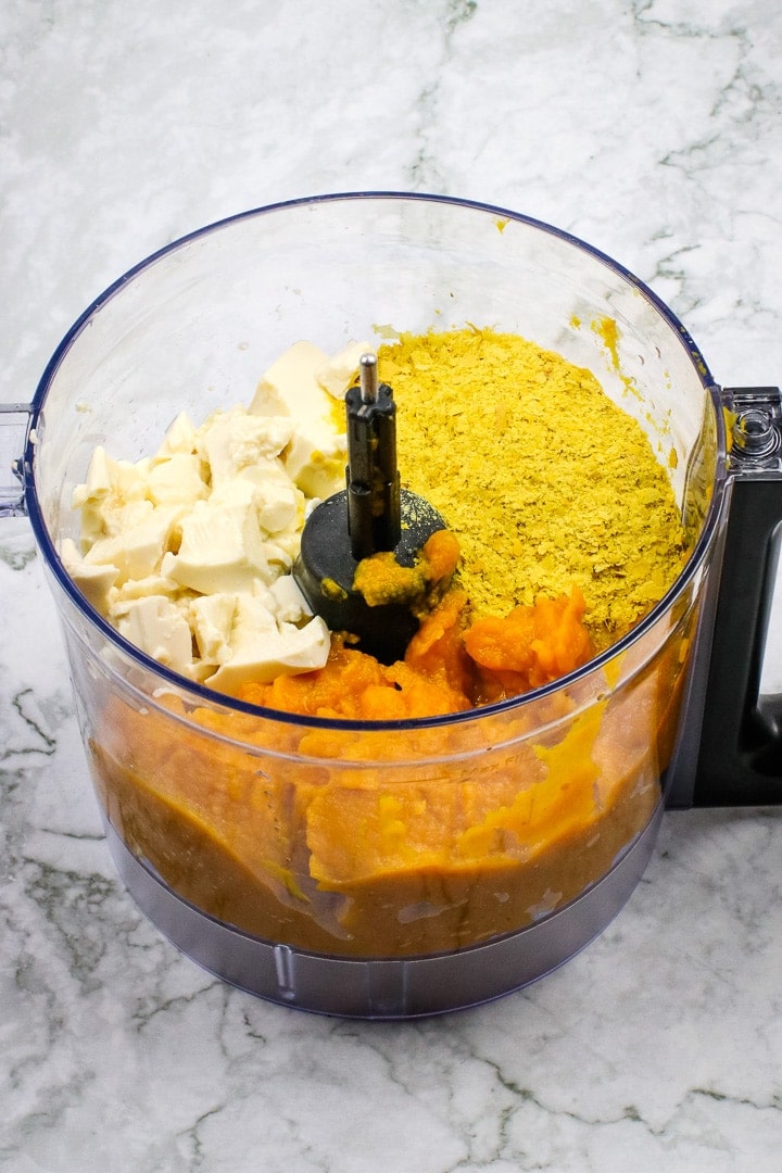 Food processor bowl with tofu, nutritional yeast and pumpkin puree on gray marble.