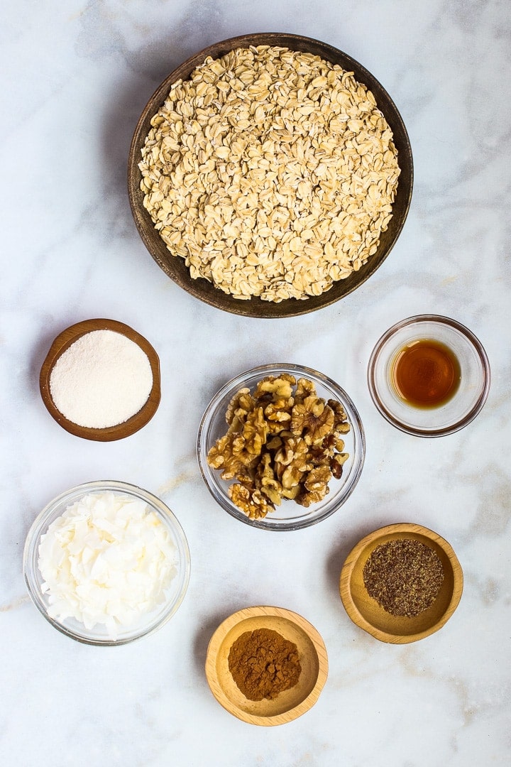 Bowls of oats, erythritol, walnuts, flax, vanilla, coconut flakes and cinnamon on white marble.