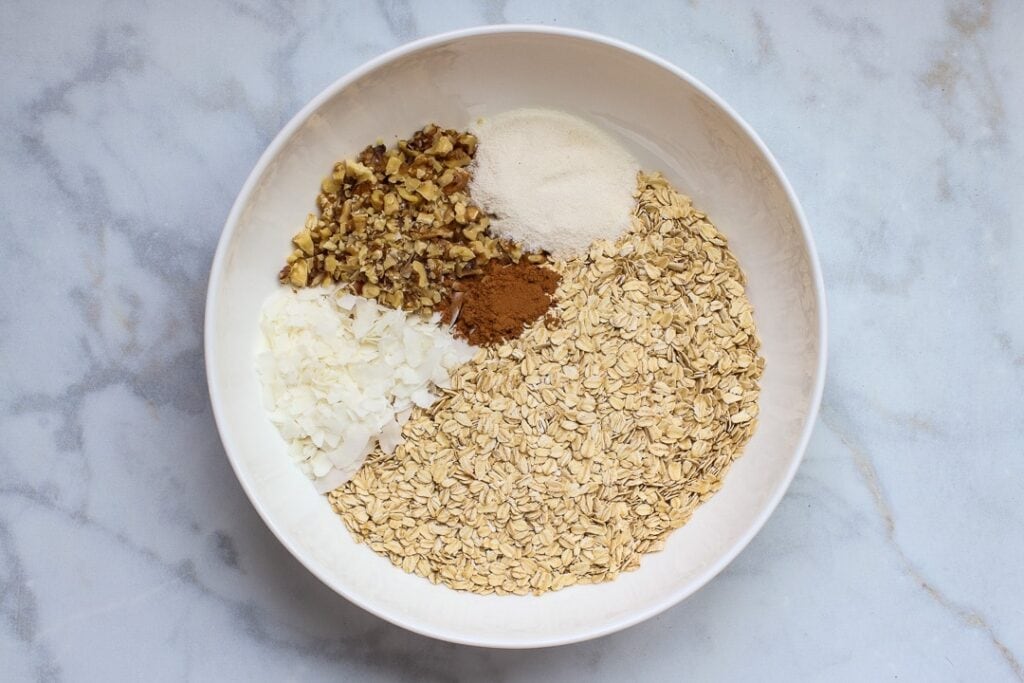 White bowl with oats, chopped nuts, coconut flakes, cinnamon and erythritol on white marble.