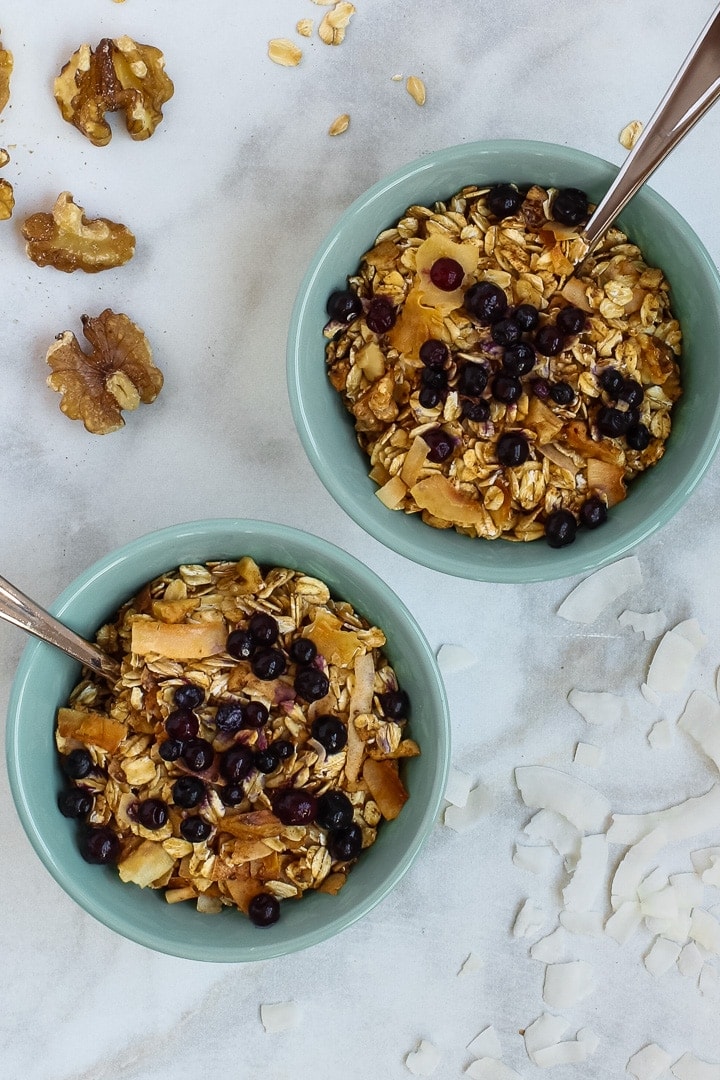 Two aqua bowls of granola with blueberries and spoons, with oats, walnuts and coconut flakes on marble.