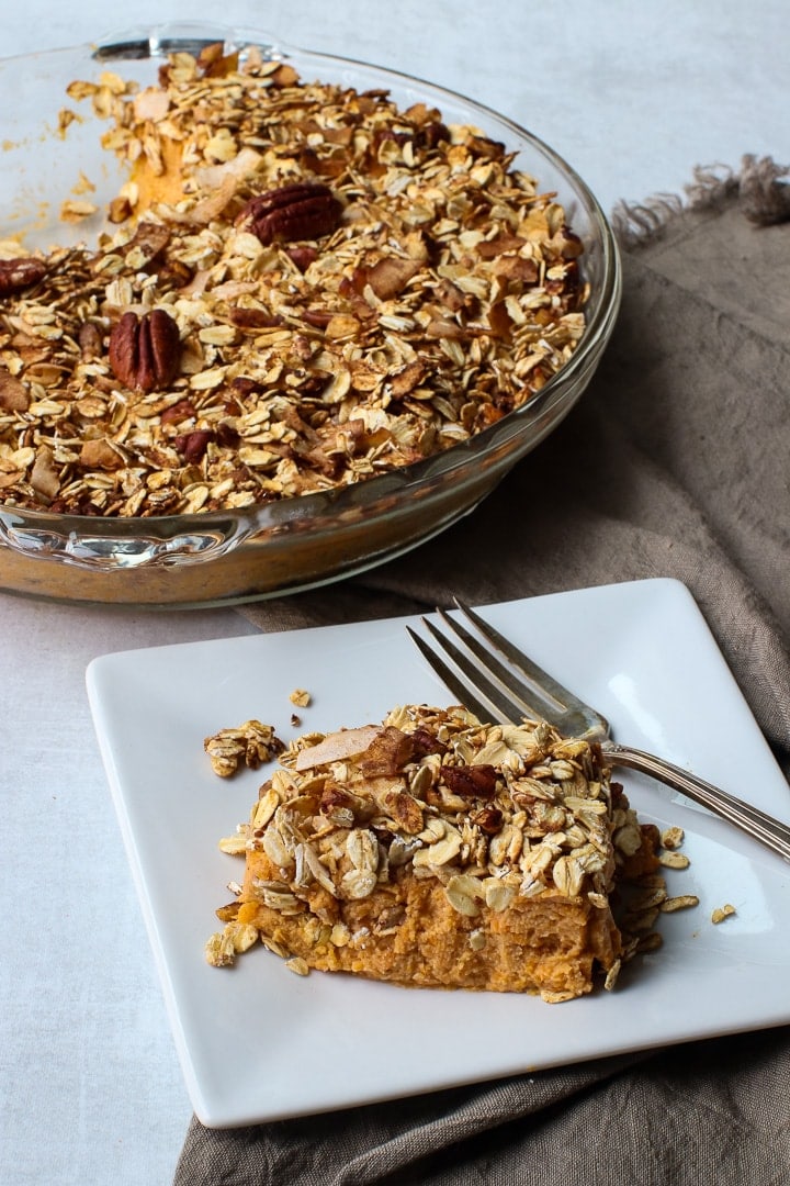 Sweet potato casserole in clear glass pie dish on brown napkin; white plate of sweet potato casserole and fork in foreground.