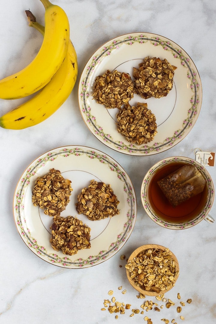 Vegan oatmeal cookies on tow antique china plates; two banana, small bowl of granola and cup of tea on white marble.