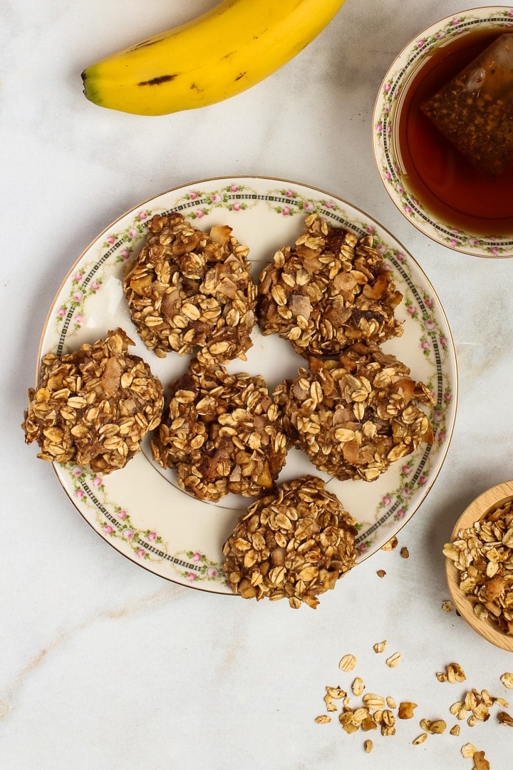 Vegan oatmeal cookies on an antique china plate; banana, small bowl of granola and cup of tea on white marble.