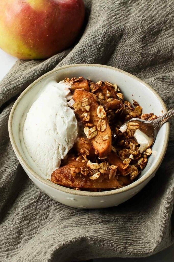 close up of apple crisp in bowl with ice cream and spoon and apple on brown towel
