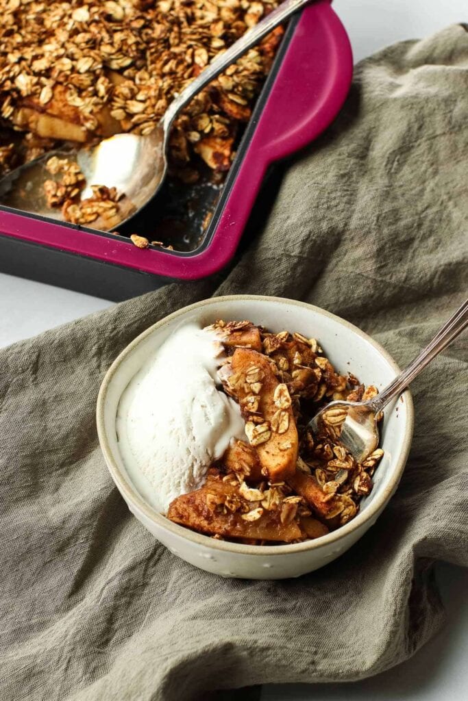 Baking dish with apple crips and spoon; small bowl of apple crips and ice cream with spoon on brown towel.