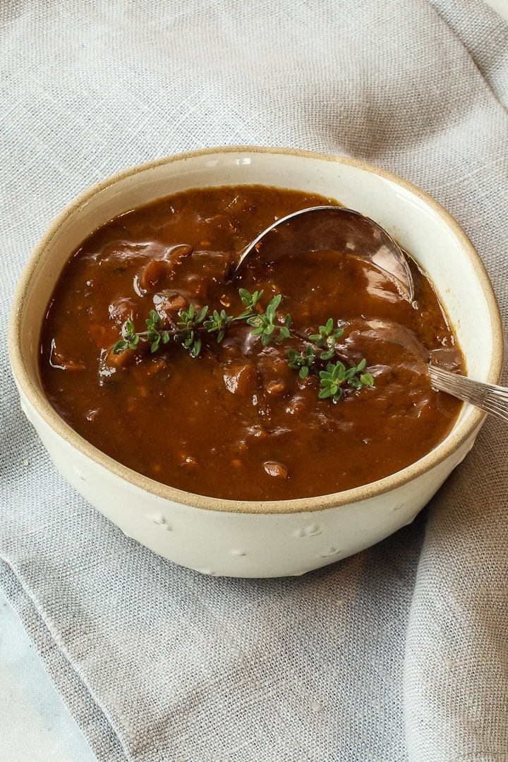 Gray bowl of porcini mushroom gravy with ladle and thyme sprig on gray-blue napkin