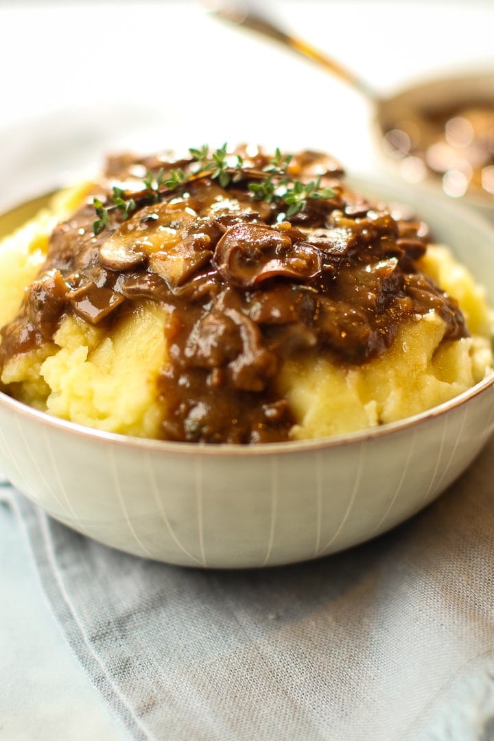 Gray bowl of mashed potatoes with glistening mushroom gravy on top, bowl and ladle in background.