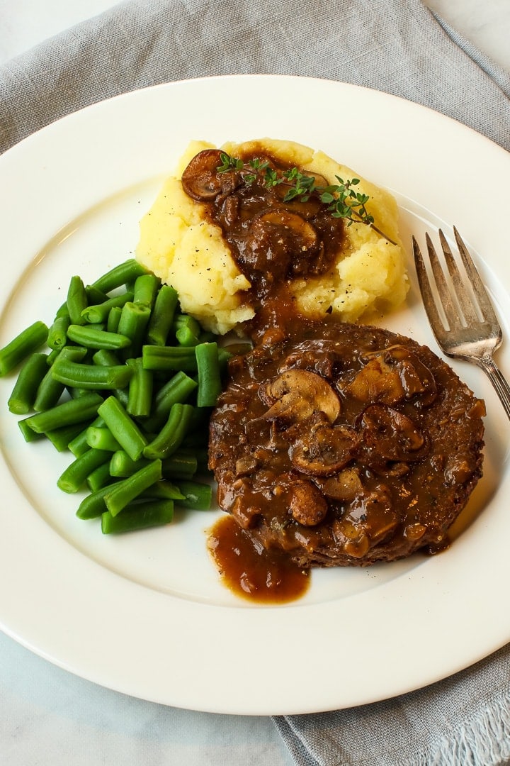 White china plate with green beans, and tofu walnut burger and mashed potatoes smothered in gravy, with fork.