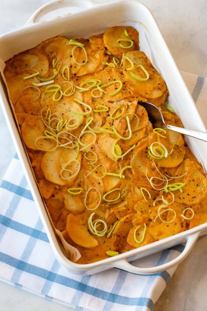 cheesy potato casserole topped with leeks in white baking dish on blue plaid towel