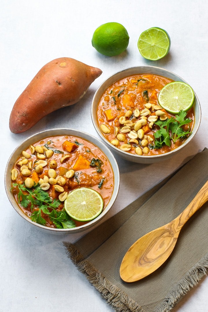 Two bowls of African peanut stew with lime slice, chopped peanuts and cilantro. Whole sweet potato , wooden spoon and cut lime in background.