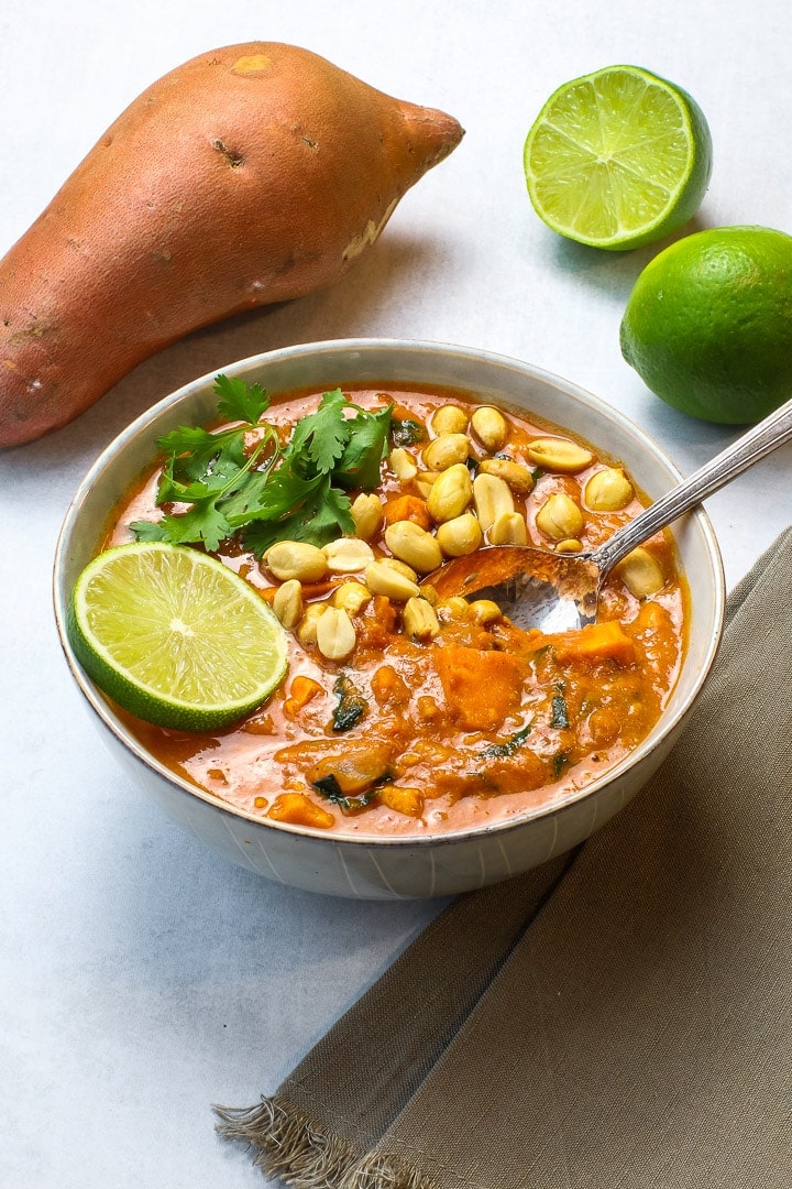 Bowl of African peanut stew with spoon, lime slice, chopped peanuts and cilantro. Whole sweet potato and cut lime in background. 
