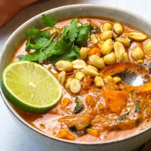 Bowl of African peanut stew with spoon, lime slice, chopped peanuts and cilantro.