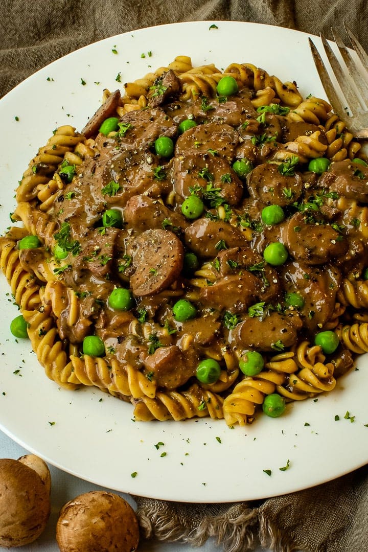 Close up of white plate with mushroom stroganoff over curly pasta, garnished with parsley, with fresh mushrooms and fork on the side.