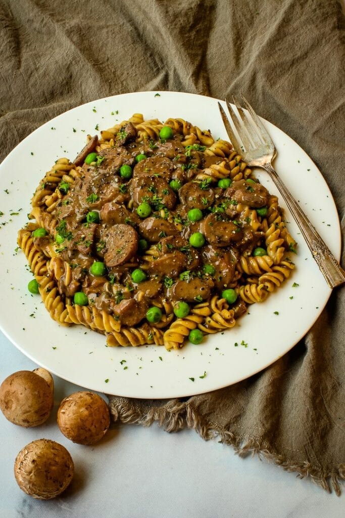 white plate with mushroom stroganoff over curly pasta, garnished with parsley, with fresh mushrooms and fork on the side.