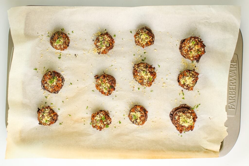 Stuffed mushrooms topped with more vegan Parmesan and chopped parsley for serving, on a parchment paper lined baking sheet.