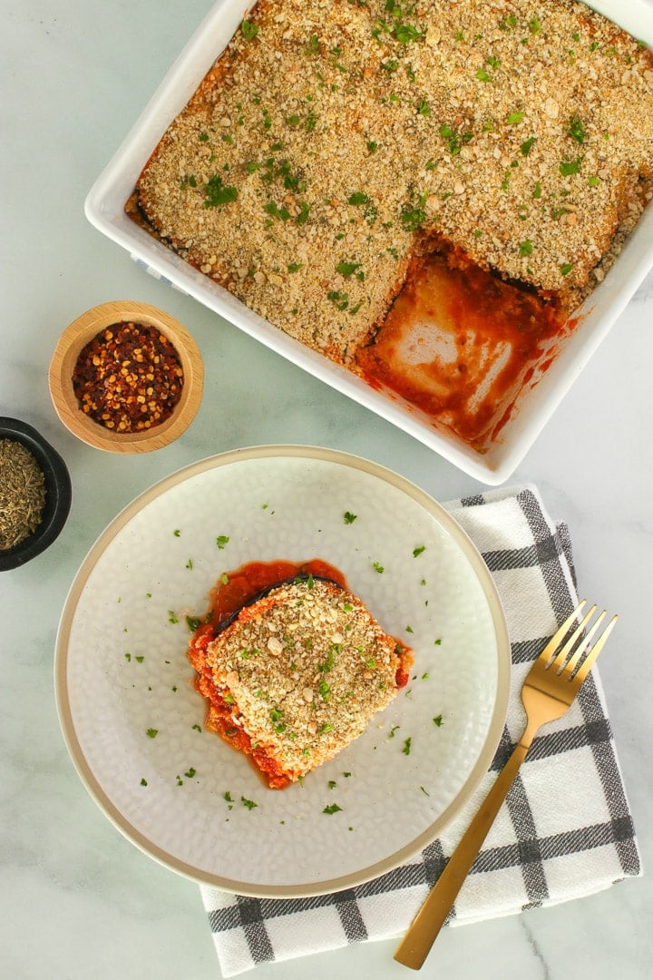 Vegan eggplant Parmesan slice on a white plate, with casserole dish in background. Gold fork, black plaid napkin and spices.