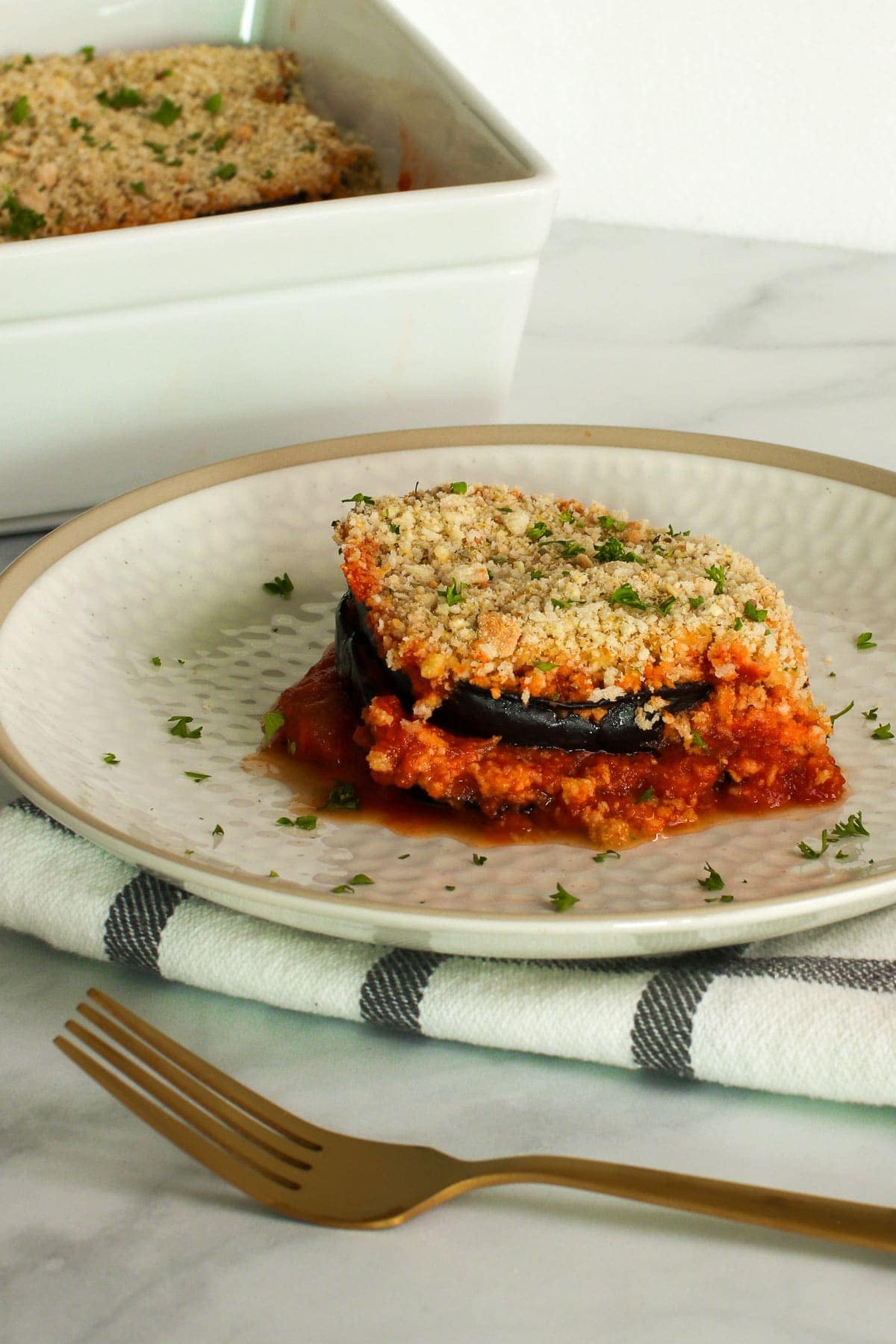 Vegan eggplant Parmesan slice on a white plate, with casserole dish in background. Gold fork, black plaid napkin.