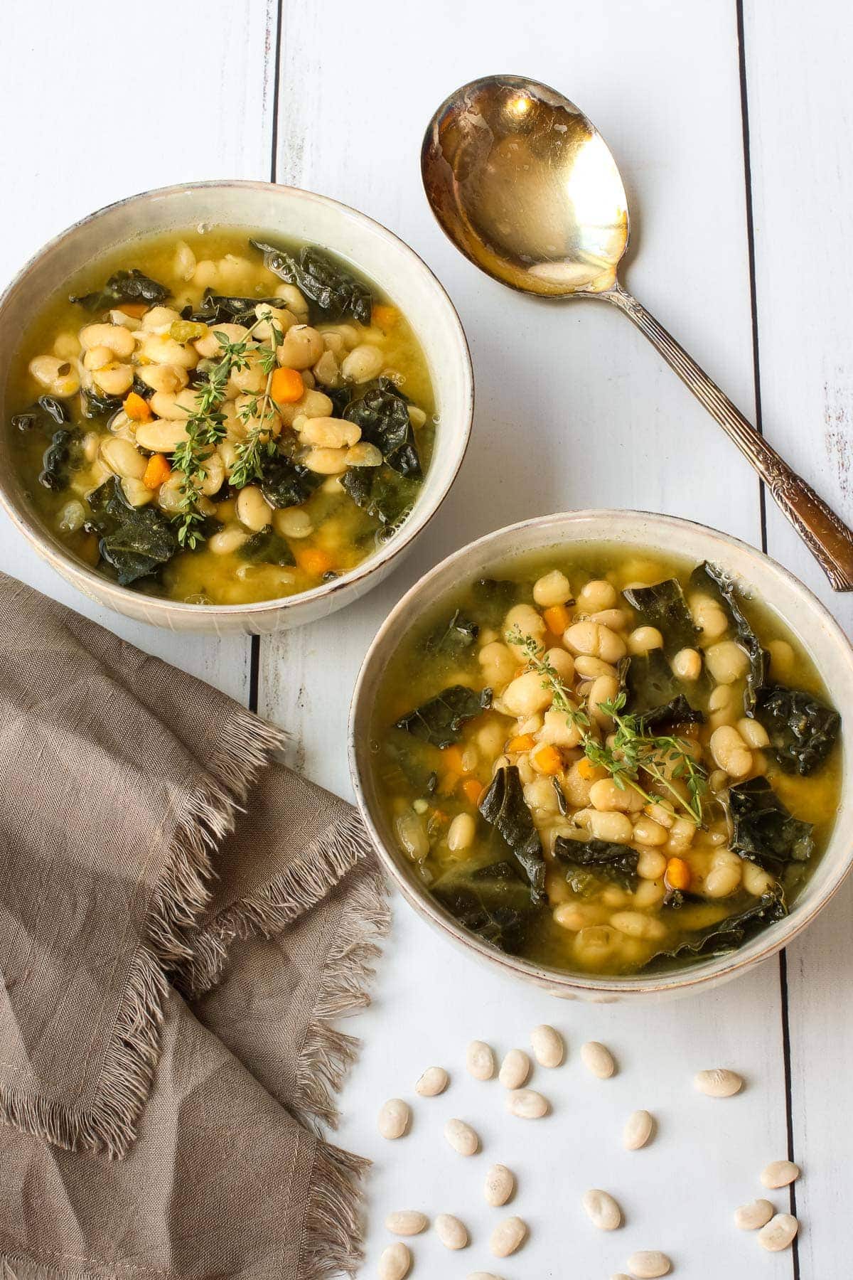 two bowls of white bean and kale soup with spoon, dried beans and brown towel. 