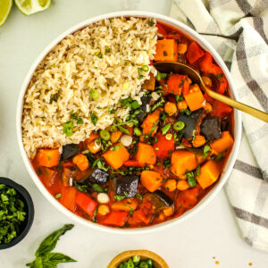 curry stew in a white bowl with rice, fresh herbs sprinkled on top, with limes, towel, gold spoon, basil leaf and small dishes of herbs.