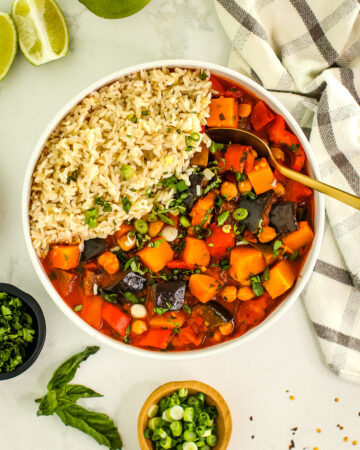 curry stew in a white bowl with rice, fresh herbs sprinkled on top, with limes, towel, gold spoon, basil leaf and small dishes of herbs.