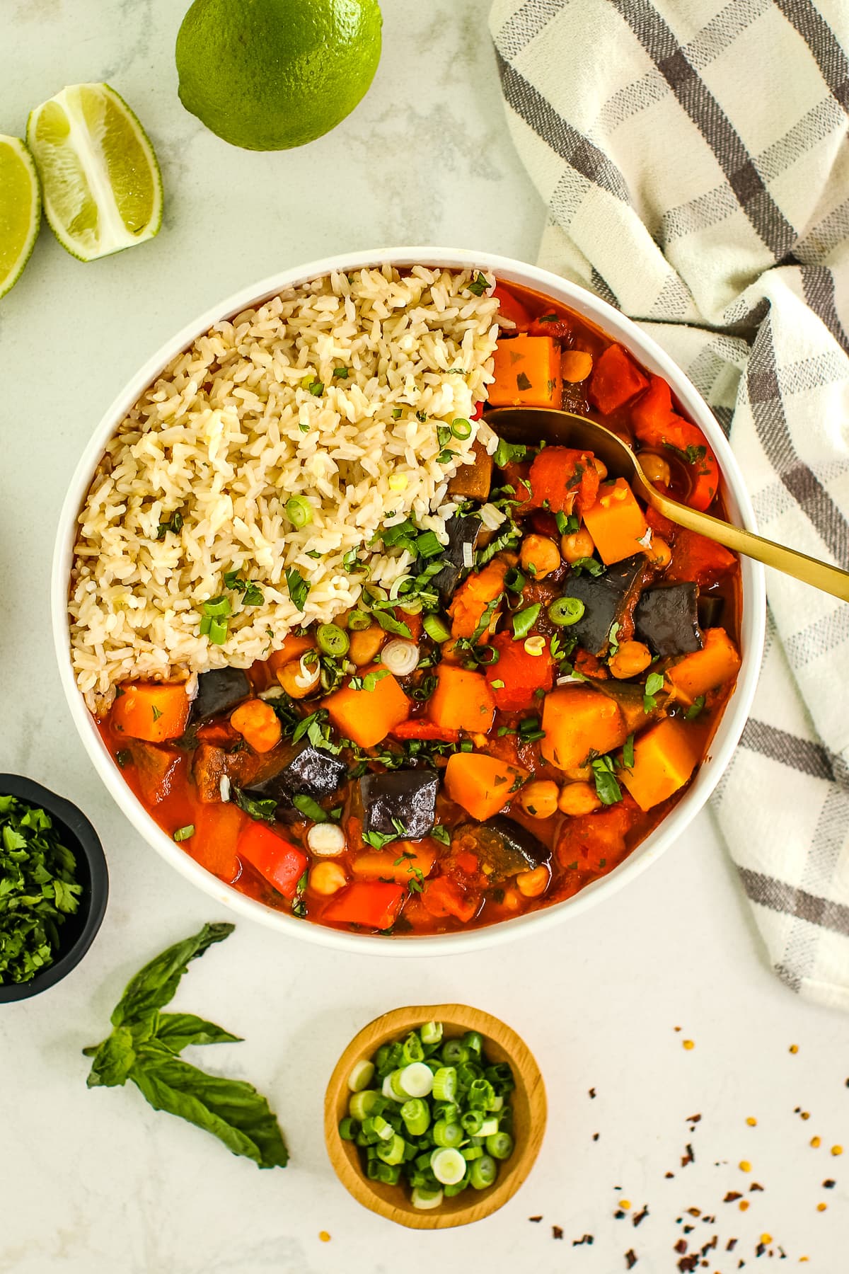 curry stew in a white bowl with rice, fresh herbs sprinkled on top, with limes, towel, gold spoon, basil leaf and small dishes of herbs.