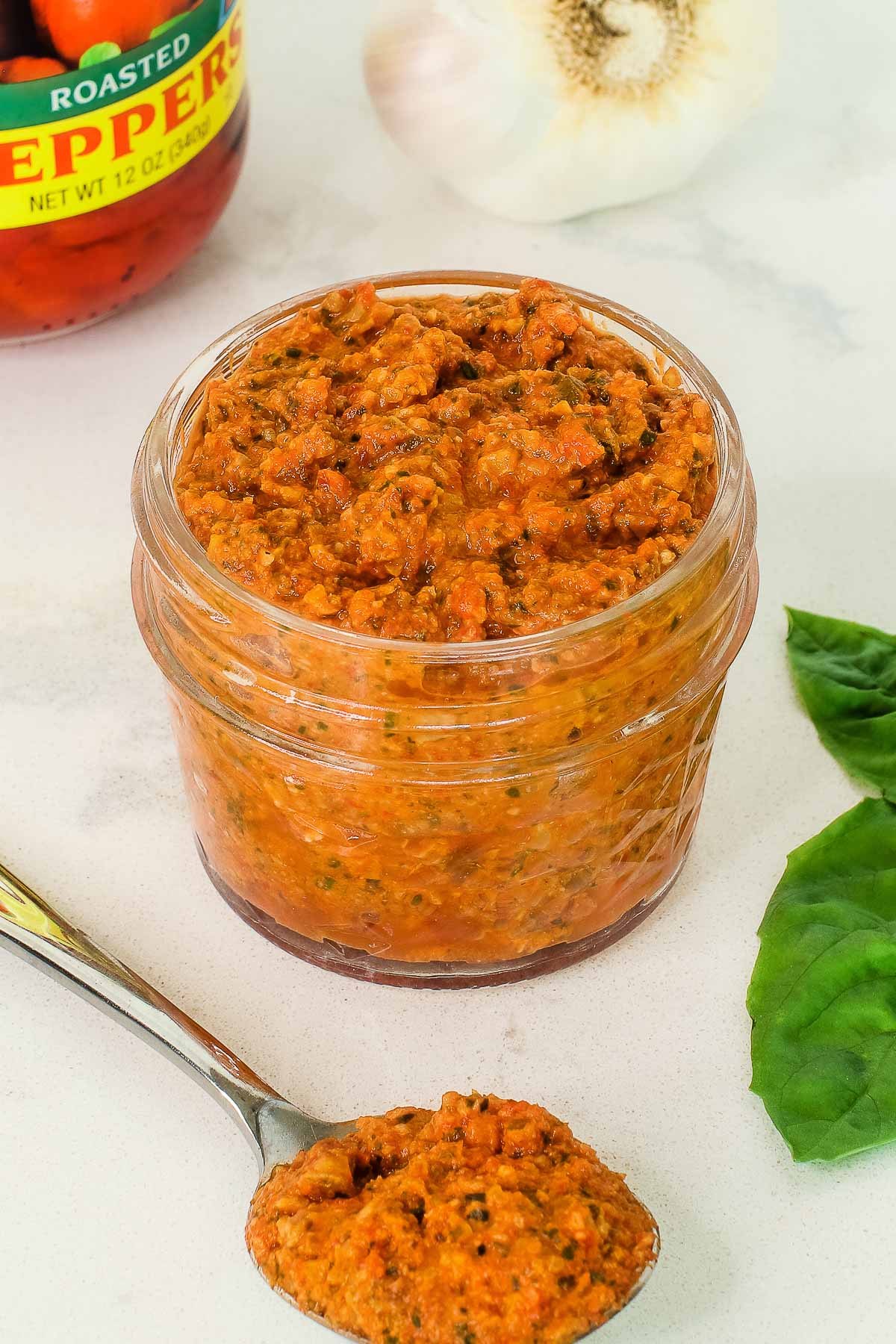 close up of jar of pesto with spoon, basil and red pepper jar and garlic in background