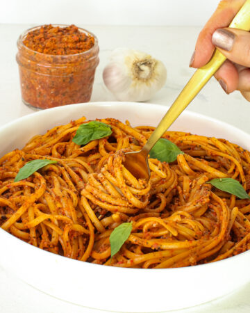 close up of red pepper pasta in bowl, fork twirling, jar of pesto and garlic in background