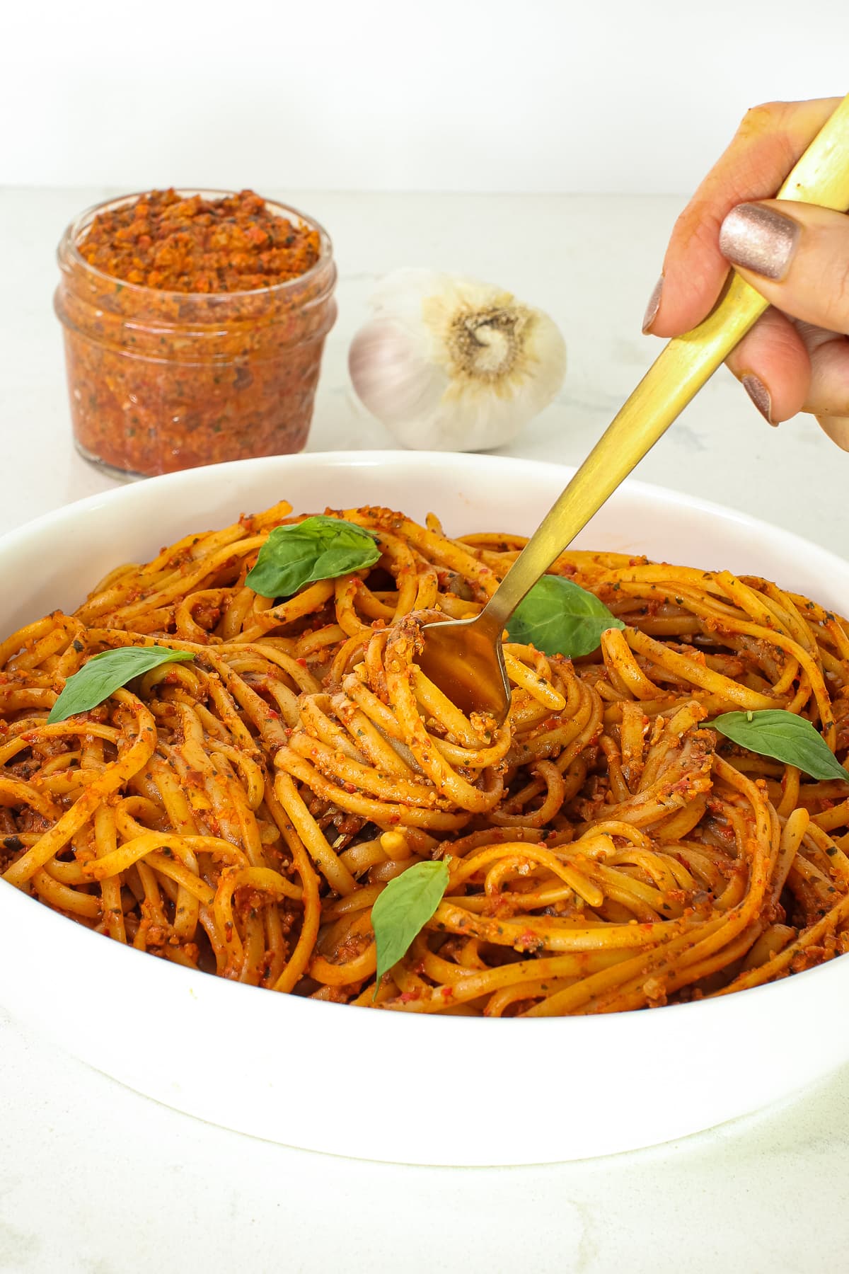 close up of red pepper pasta in bowl, fork twirling, jar of pesto and garlic in background