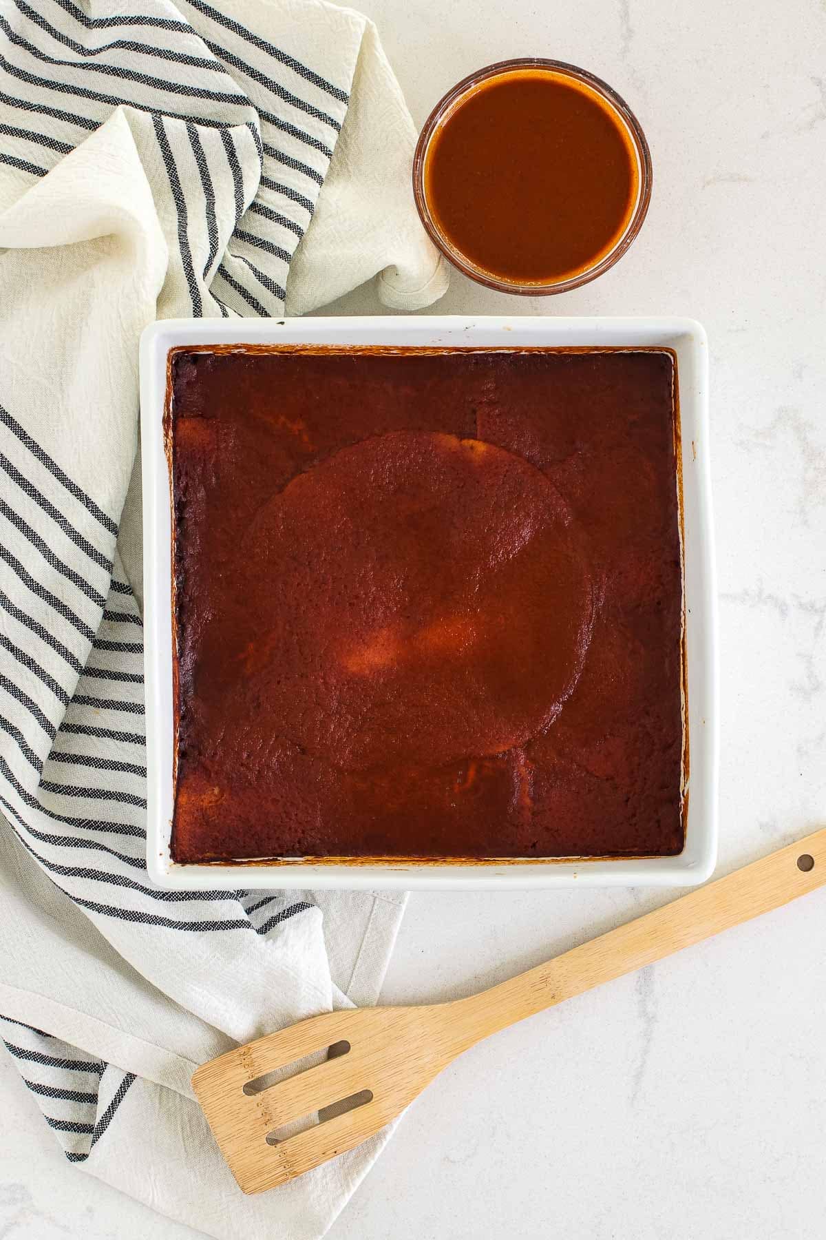 baked casserole in square dish with towel, extra sauce and wooden spoon.