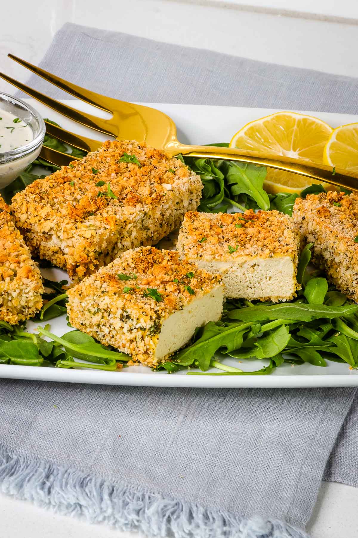 Close up of cut baked tofu chicken breasts on a platter with arugula, lemon and a dish of ranch dressing. 