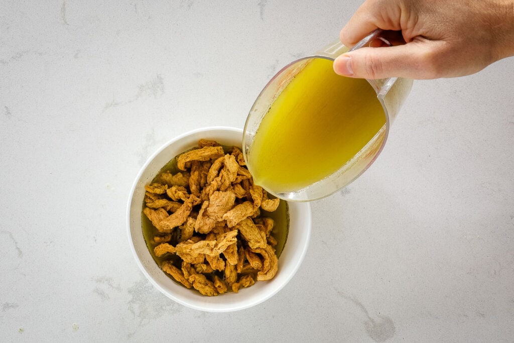 Pouring broth from a measuring cup over a bowl of soy curls.
