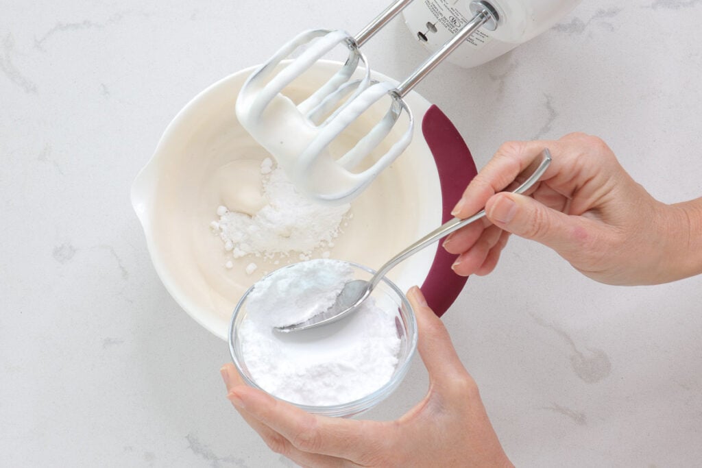 Spooning powdered sugar into the bowl, beaters covered in topping. 